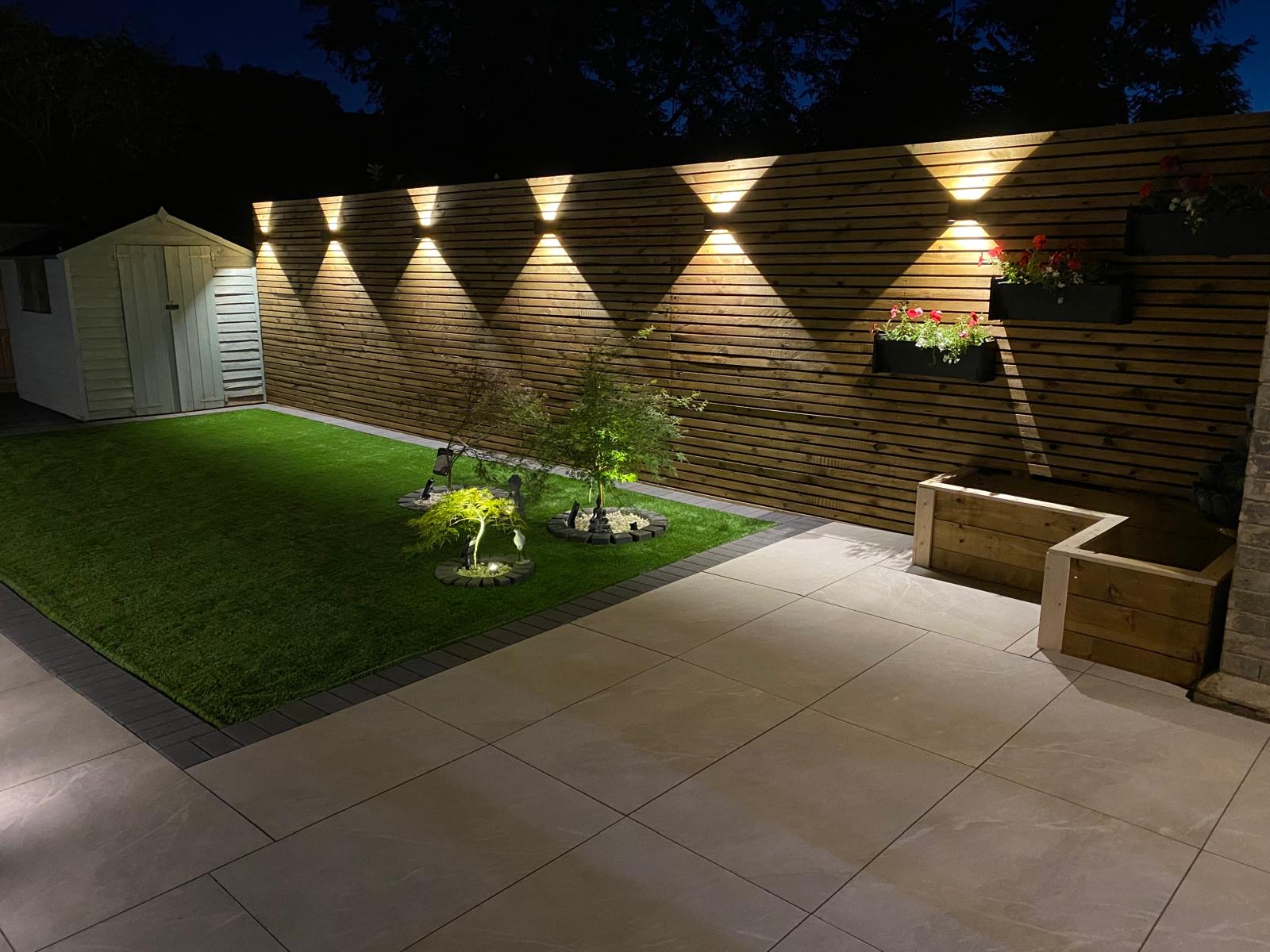 Slatted cedar fence with wall lighting behind a porcelain patio and artificial lawn, built by Golden Hands